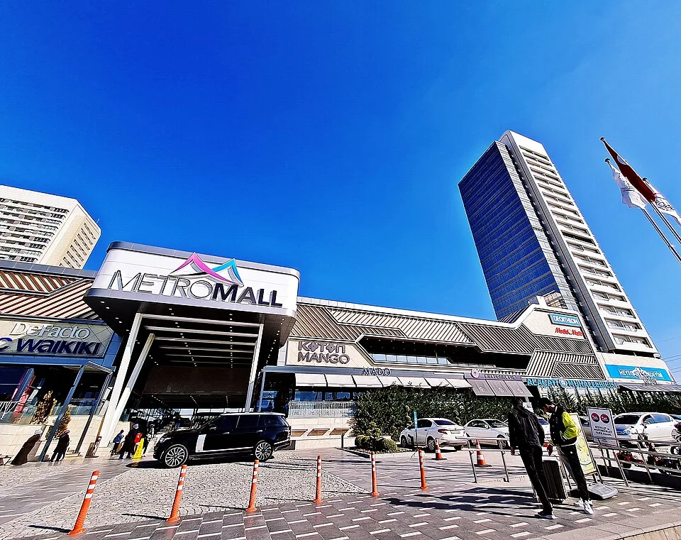 Metromall AVM'nin tabelalı ana girişi, önündeki araçlar ve arkasındaki yüksek yapılar