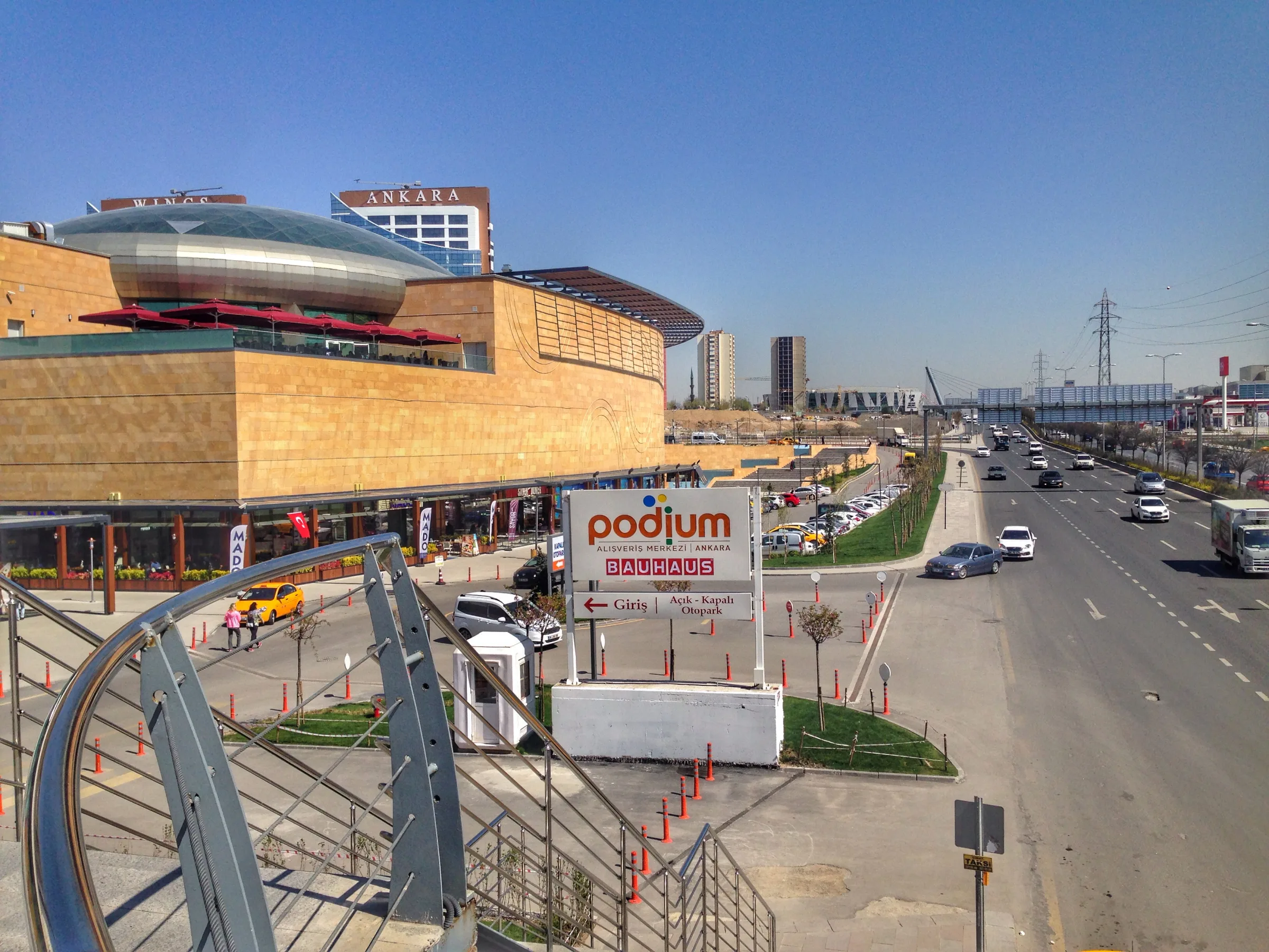 Curved stone-clad exterior of Ankara Podium shopping centre beside its entrance sign, outdoor stairs and Bağdat Avenue